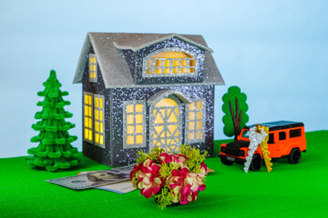 On a light background with yellow light in the windows, a gray house surrounded by an orange car with keys and euro and dollar banknotes