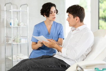 Obraz premium Young male patient lying at examination couch reading and signing documents while talking with female doctor