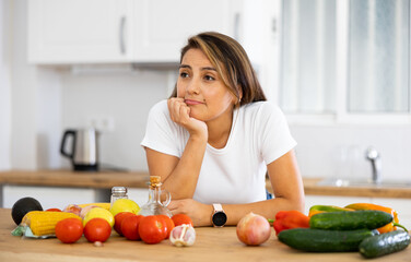 Portrait of upset frustrated young Hispanic woman standing in home kitchen, leaning on table full of fresh vegetables