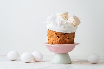 Easter cake decorated with white icing and macaroons. Traditional sweet Easter bread on the stand surrounded with colorful painted eggs on white background. Copy space