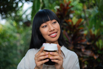 Smiling Asian woman holding a reusable coffee cup in a lush green garden.