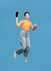 Excited woman jumping with phone in hand giving thumbs up on blue background Victory gesture