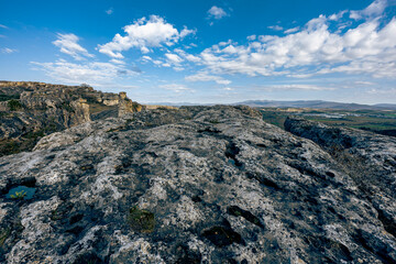 Wide view from a rocky plateau with dramatic textures, scattered clouds in a vivid blue sky, and distant green fields under mountain foothills.