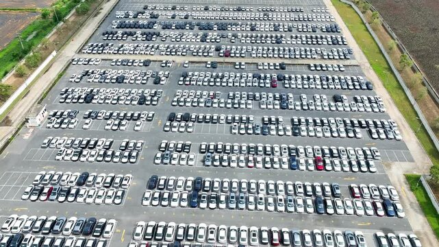 The aerial perspective offers a striking view of a large parking lot, where a sea of white cars stands in perfect rows, with a few scattered red and black vehicles offering contrast.
