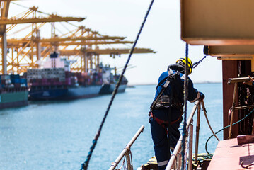 A deckhand prepares the pilot ladder as the ship readies for port entry. Secured in a safety harness, he carefully ensures the setup is ready, focusing on the task in a high-risk environment. 