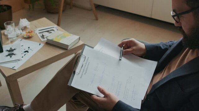 High angle shot of male bearded psychologist in smart casual outfit and glasses filling out mental health assessment forms during therapy session, changing papers in clipboard