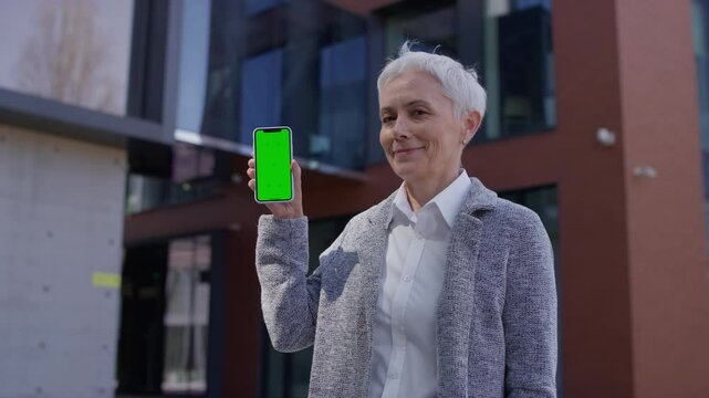 Smiling senior woman showing smartphone with green screen outdoors