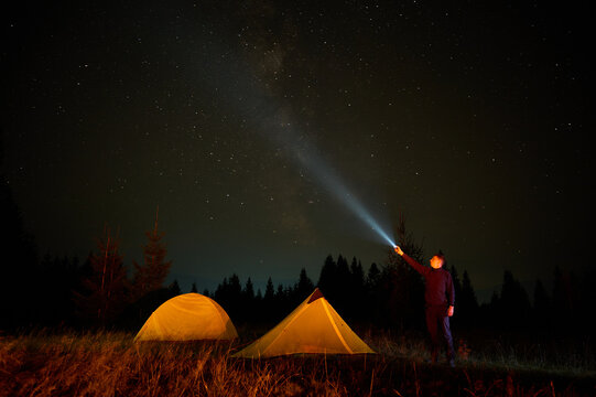 Man tourist with flashlight near his camp ten. Camping tents under the starry sky.  Orange illuminated tent. iluminated tent under stars in the mountains