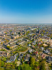Fototapeta premium Aerial view of a Dutch town with a mix of modern and traditional houses, historic churches, and distant farmlands under a clear blue sky on a sunny spring day.