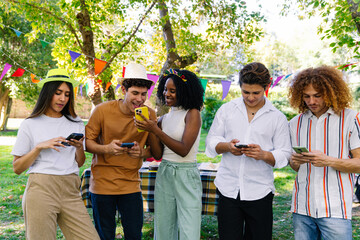 Five young friends using smartphones during a birthday party in a park, celebrating and sharing moments