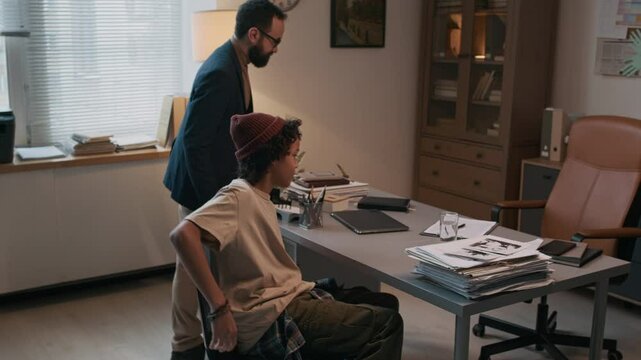 Tracking shot schoolboy in casual clothes and beanie entering office of male school psychologist, boy sitting on chair and casually putting bag on floor, having anxiety problems or being bullied