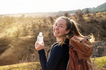 Young woman drinking water from a plastic bottle while halt, looking at the camera with a friendly...