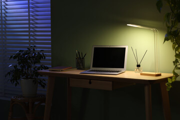 Comfortable workplace with laptop and lamp on wooden desk at home office in evening
