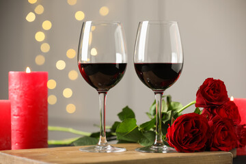 Glasses of red wine, burning candle and roses on table against gray background with blurred lights. Romantic dinner