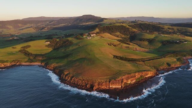 Aerial orbit of vibrant green fields and ocean cliffs near Kiama coastline under clear skies, Australia