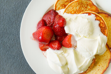 Close up of home made pancakes with strawberries  and whipped cream on a white plate with grey felt background, flat lay