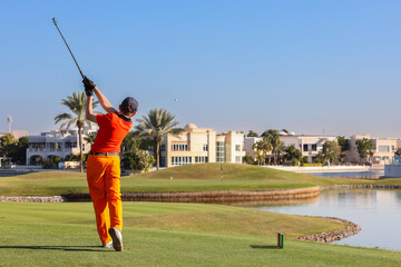 Golfer taking swing on luxury waterfront course, Male golfer in bright outfit swinging club on a high-end golf course near water.