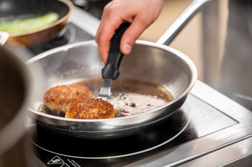 A close-up of a hand using a spatula to flip two golden-brown patties in a sizzling skillet, illustrating the cooking process in a professional kitchen environment.