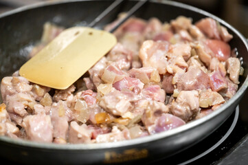 A close-up of chopped chicken pieces sizzling in a frying pan, with a spatula resting on the side, depicting the preparation of a savory dish in a kitchen.