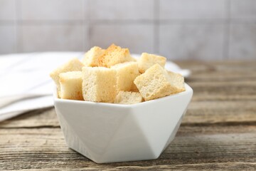 Tasty crispy croutons in bowl on wooden table against grey tiled wall, closeup