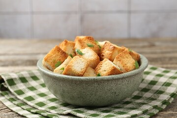 Tasty crispy croutons with cut parsley in bowl on wooden table against grey tiled wall, closeup