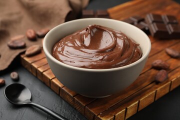 Tasty pudding in bowl, pieces of chocolate and cocoa beans on black table, closeup
