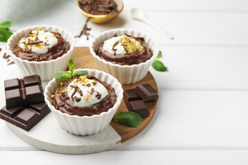 Tasty pudding with whipped cream in bowls and pieces of chocolate on white wooden table, space for text