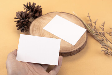 Hand holding white blank business card mockup with wooden decoration and dried flower on brown background
