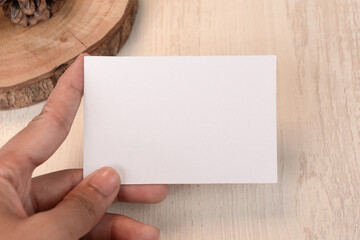 Obraz premium Hand holding white blank business card mockup with wooden decoration on brown background