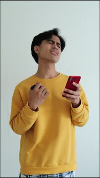 Disappointed young Indonesian Asian man holding a credit card while looking at smartphone, expressing frustration or regret, isolated on white background in 4K resolution.