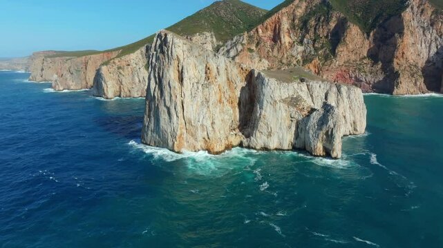 Low drone establishing shot of Pan di Zucchero sea stack near Masua, Sardinia, the limestone monolith rises sharply from vivid blue waters, surrounded by dramatic cliffs and lush hills