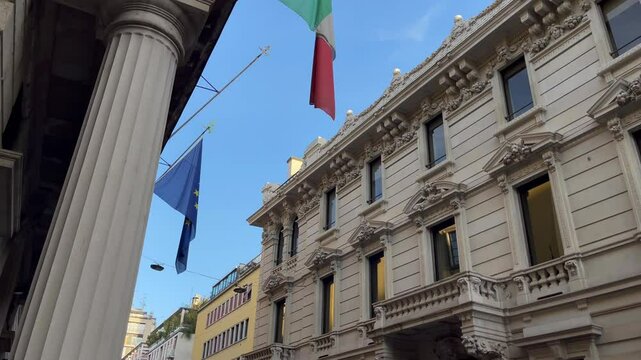 Ornate historic buildings in Milan with neoclassical facades and Italian and EU flags waving under a clear blue sky, viewed from street level