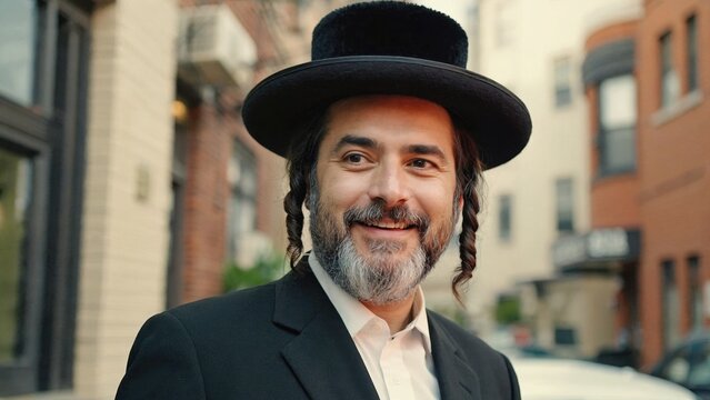 A happy, bearded Jewish man wearing a black yamaka and hat on city street