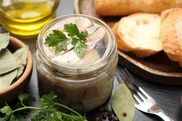 Pieces of delicious herring with spices in jar and bread on black table, closeup