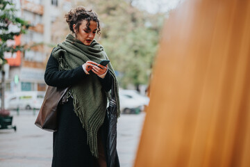 A stylish woman with a scarf browses her phone while walking in an urban environment.