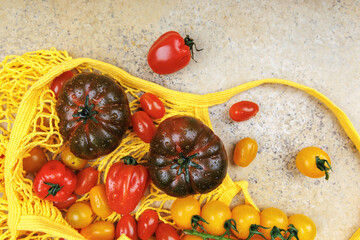 Top view of fresh heirloom and cherry tomatoes in yellow mesh grocery bag on stone background