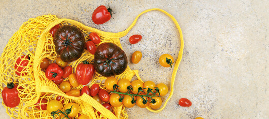 Assorted fresh tomatoes in yellow reusable mesh shopping bag on stone surface