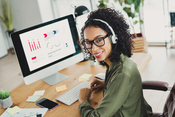 Young professional woman wearing headphones working in a modern office setting, analyzing data charts on a large computer monitor