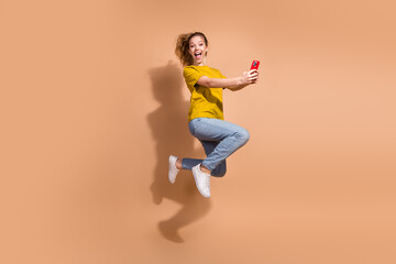 Young woman enthusiastically jumping in casual attire with smartphone against beige backdrop