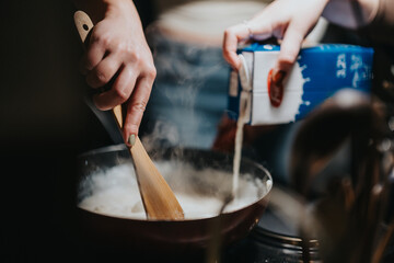 A detailed view of two hands cooking, one stirring with a wooden spoon and the other pouring milk into a saucepan, exuding a homely culinary atmosphere.
