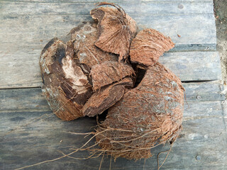 A cracked, dried coconut shell with fibrous husk remnants, resting on a rustic wooden surface. The shell appears weathered, likely used or naturally aged.