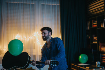 A man enthusiastically plays guitar amidst a festive home setting with balloons and lights,...