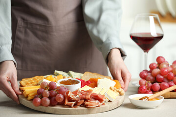 Woman holding board with different types of cut cheese and other snacks at light textured table, closeup