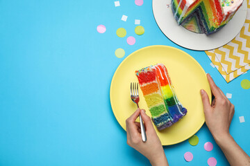 Woman eating delicious rainbow cake at light blue table with confetti, top view. Space for text
