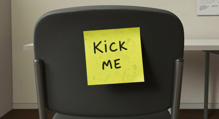 Humorous sticky note reading "Kick Me" attached to the back of a chair in an office setting, creating a lighthearted prank atmosphere

