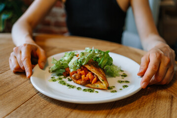 Woman's hands holding a plate with a folded omelette filled with chickpeas, carrots, salad and pesto sauce, served on a wooden table, suggesting a healthy and delicious breakfast or brunch option