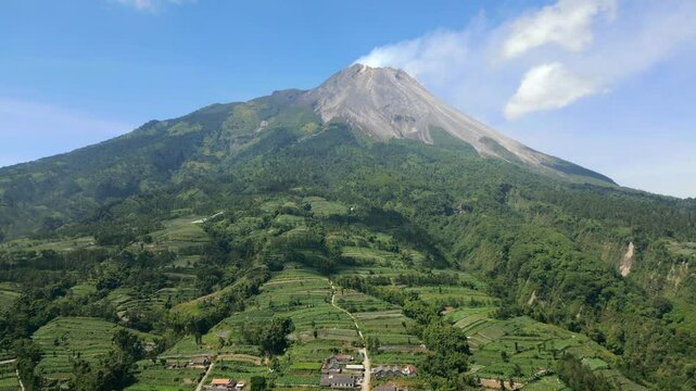 Aerial view of Mount Merapi volcano surrounded by lush green farmland and rural landscape in Java, Indonesia. Scenic volcanic terrain and cultivated hills under clear blue sky.