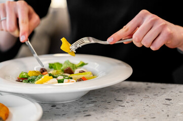 A close-up of hands using a fork and knife to enjoy a fresh salad, featuring colorful vegetables and greens, highlighting the dining experience in a cozy setting.