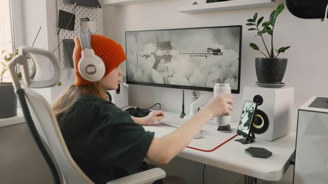 Teen gamer girl in orange beanie sits in ergonomic chair and plays online shooter on widescreen monitor. Clean setup, immersive play and modern digital lifestyle of Gen Z. Woman drinking energy drink 