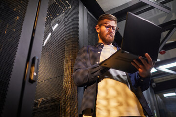 Man inspecting server rack while holding laptop in modern data center with focused expression. Image shows male professional in technology-rich setting